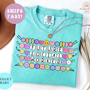 May include: A turquoise t-shirt with the words "FUTURE SOCIAL WORKER" in white letters, surrounded by colorful beads and decorative elements. The shirt has a crew neck and short sleeves. A gold necklace is draped across the shirt.