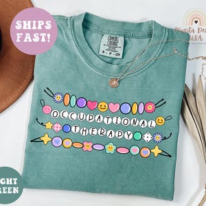 May include: Light green t-shirt with the words "OCCUPATIONAL THERAPY" in white letters, decorated with colorful beads and flower graphics. The shirt has a crew neck and short sleeves. A necklace is draped across the shirt.