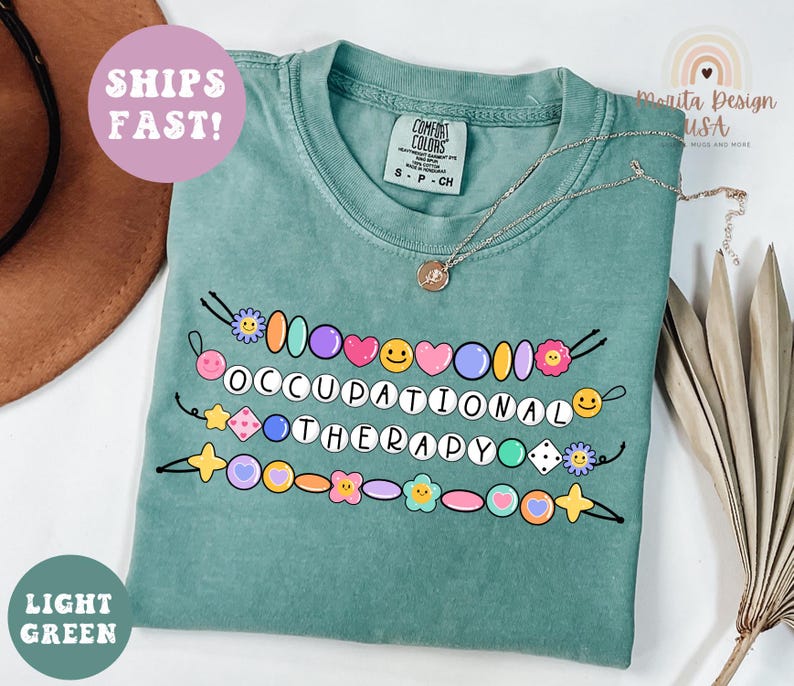 May include: Light green t-shirt with the words "OCCUPATIONAL THERAPY" in white letters, decorated with colorful beads and flower graphics. The shirt has a crew neck and short sleeves. A necklace is draped across the shirt.