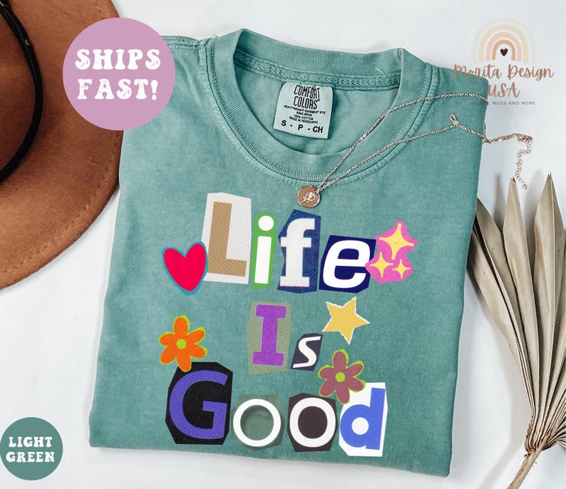 May include: A light green t-shirt with the phrase "Life Is Good" in a collage style. The design includes a red heart, stars, and flowers. A silver necklace with a pendant is on the shirt.