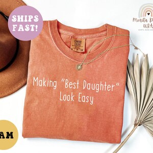 May include: A coral-colored t-shirt with the phrase "Making 'Best Daughter' Look Easy" in white. A gold necklace and a brown hat are visible. The shirt has a Comfort Colors tag.