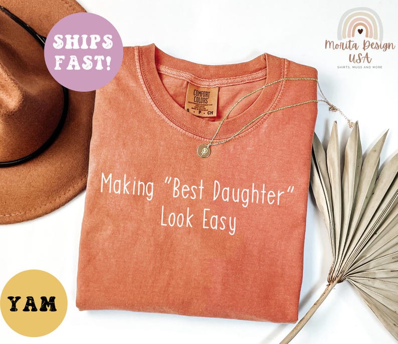 May include: A coral-colored t-shirt with the phrase "Making 'Best Daughter' Look Easy" in white. A gold necklace and a brown hat are visible. The shirt has a Comfort Colors tag.