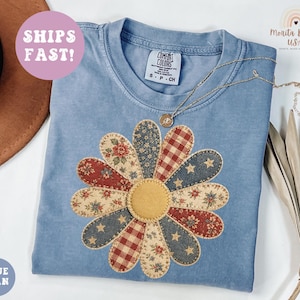 May include: A light blue, short-sleeved t-shirt with a large, appliqued daisy design. The daisy petals feature floral, star, and plaid patterns in red, blue, and cream. A gold necklace rests on the shirt. The text "BLUE JEAN" is in a blue circle.