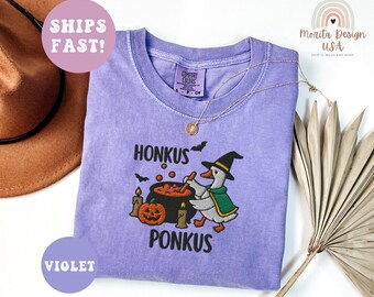 May include: A violet-colored t-shirt with the embroidered text "HONKUS PONKUS" and a cartoon goose wearing a witch hat and cape, stirring a cauldron. The design includes a jack-o'-lantern and candles. A gold necklace is draped on the shirt.