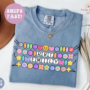 May include: Blue jean colored t-shirt with a colorful beaded design spelling "EMILY". The design includes various shapes like hearts, stars, and flowers. The shirt also has the text "SHIPS FAST!" and "BLUE JEAN".