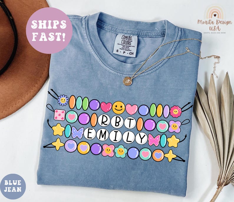 May include: Blue jean colored t-shirt with a colorful beaded design spelling "EMILY". The design includes various shapes like hearts, stars, and flowers. The shirt also has the text "SHIPS FAST!" and "BLUE JEAN".