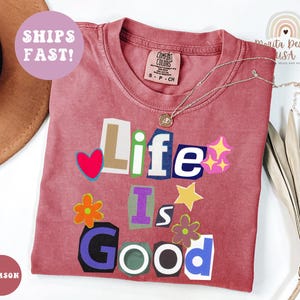 May include: Crimson-colored t-shirt with the phrase "Life Is Good" in a colorful, cut-out style. The shirt features a crew neck and a small tag. A silver necklace with a pendant is draped across the shirt.