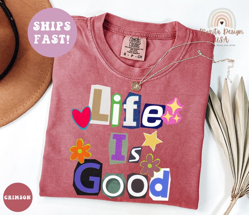 May include: Crimson-colored t-shirt with the phrase "Life Is Good" in a colorful, cut-out style. The shirt features a crew neck and a small tag. A silver necklace with a pendant is draped across the shirt.