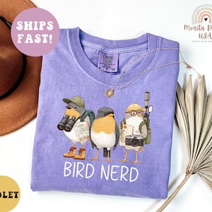 Op de afbeelding: Een paars T-shirt met een afbeelding van drie cartoonvogels, elk met een verrekijker, camera en rugzak. De tekst "BIRD NERD" staat eronder. Het shirt heeft een ronde hals en korte mouwen. Comfort Colors merk.