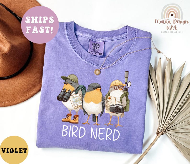 Op de afbeelding: Een paars T-shirt met een afbeelding van drie cartoonvogels, elk met een verrekijker, camera en rugzak. De tekst "BIRD NERD" staat eronder. Het shirt heeft een ronde hals en korte mouwen. Comfort Colors merk.