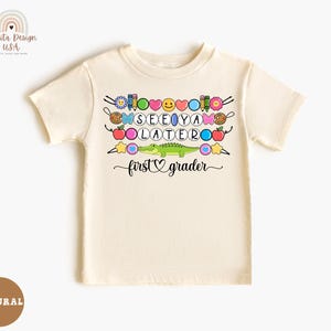 May include: Cream-colored short-sleeved t-shirt with a colorful graphic. The graphic includes the words "SEE YA LATER" and "first grader" with various school-themed and emoji-style illustrations, including a green alligator.