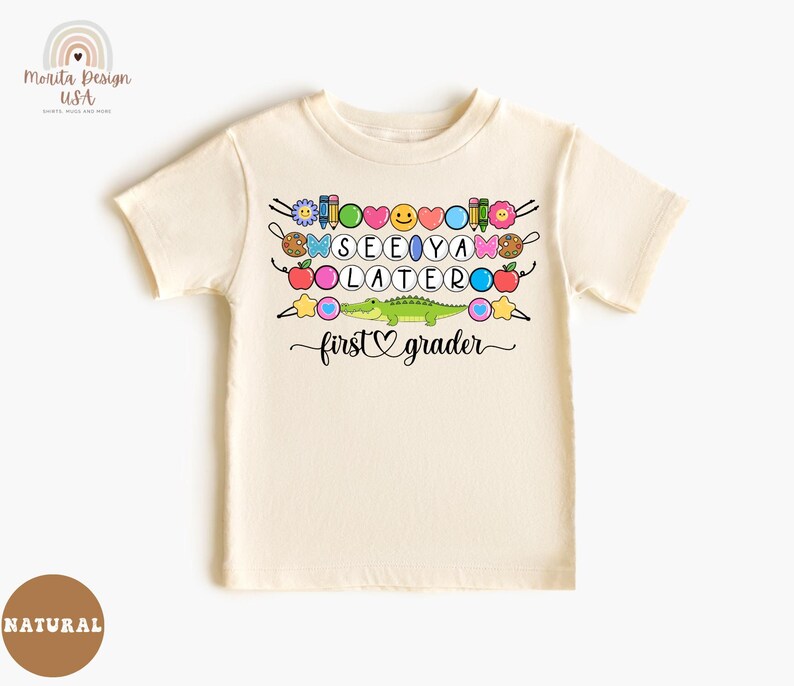 May include: Cream-colored short-sleeved t-shirt with a colorful graphic. The graphic includes the words "SEE YA LATER" and "first grader" with various school-themed and emoji-style illustrations, including a green alligator.