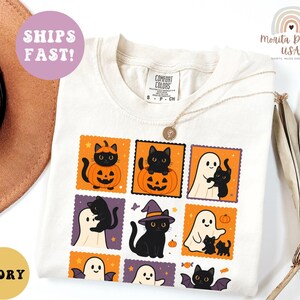 May include: Ivory-colored t-shirt featuring a Halloween-themed design. The design includes black cats, ghosts, pumpkins, and bats in a postage stamp style. The shirt also has the text "Comfort Colors" and "S P CH".