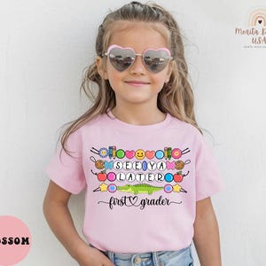 May include: A light pink t-shirt with the words "SEE YA LATERO" and "first grader" in black script. The shirt features colorful graphics of school supplies, a smiling face, and a green alligator.