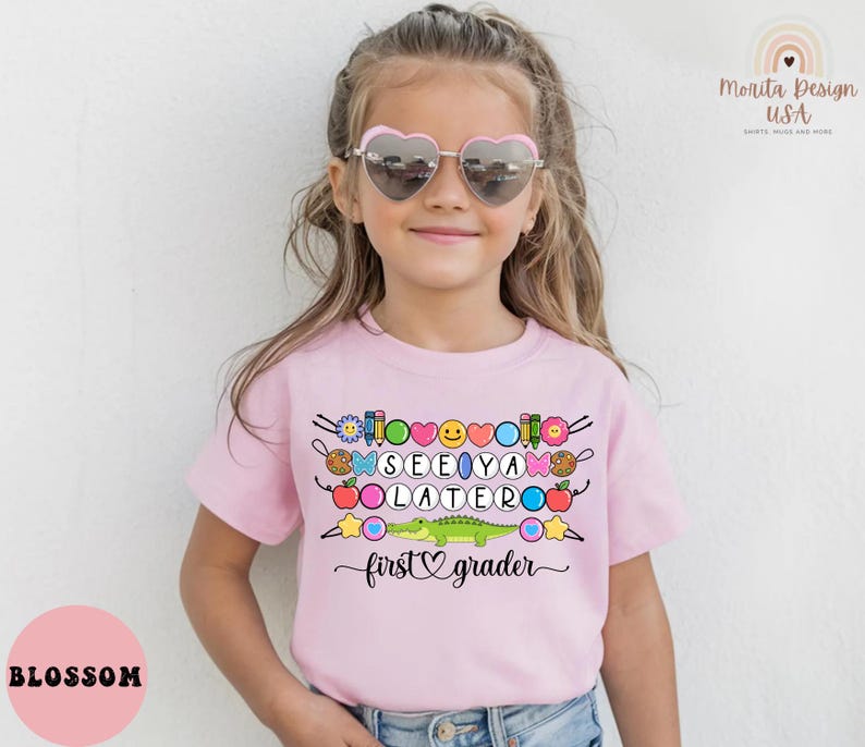 May include: A light pink t-shirt with the words "SEE YA LATERO" and "first grader" in black script. The shirt features colorful graphics of school supplies, a smiling face, and a green alligator.