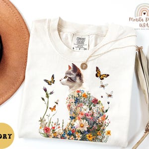 May include: A white t-shirt featuring a floral cat design. The cat is surrounded by colourful flowers and butterflies. The shirt has a tag that says "Comfort Colors" and the text "Ivory" is visible in the bottom left corner of the image.