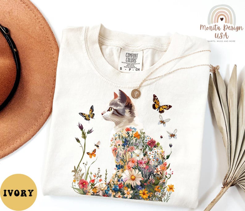 May include: A white t-shirt featuring a floral cat design. The cat is surrounded by colourful flowers and butterflies. The shirt has a tag that says "Comfort Colors" and the text "Ivory" is visible in the bottom left corner of the image.