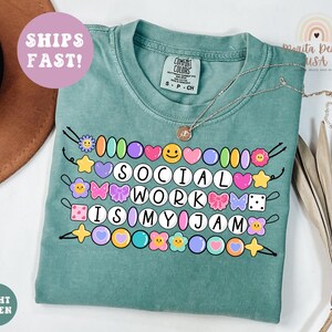 May include: A light green t-shirt with the phrase "SOCIAL WORK IS MY JAM" in a colorful, beaded design. The shirt has a crew neck and short sleeves. A silver necklace with a pendant is draped across the shirt.