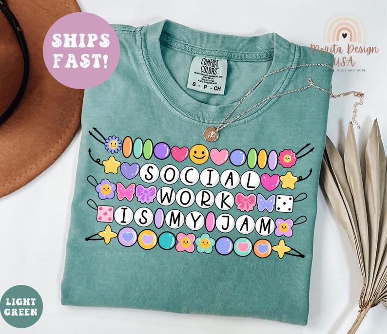 May include: A light green t-shirt with the phrase "SOCIAL WORK IS MY JAM" in a colorful, beaded design. The shirt has a crew neck and short sleeves. A silver necklace with a pendant is draped across the shirt.