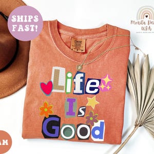May include: A coral-colored t-shirt with the phrase "Life Is Good" in a collage style. The design includes a red heart, stars, and flowers. A gold necklace and a brown hat are also visible.