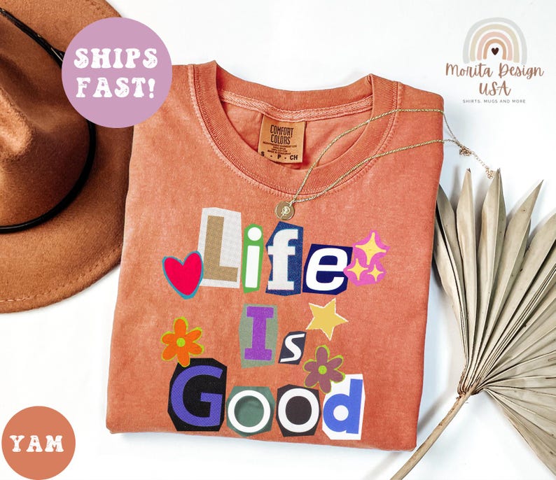 May include: A coral-colored t-shirt with the phrase "Life Is Good" in a collage style. The design includes a red heart, stars, and flowers. A gold necklace and a brown hat are also visible.