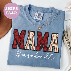 May include: A pale blue t-shirt featuring the word "MAMA" in red and cream baseball-style lettering. The word "Baseball" is written below in a cursive font. A gold necklace is draped across the shirt.