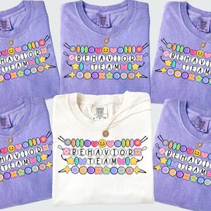 May include: A collection of lavender and white t-shirts with the text "BEHAVIOR TEAM" in a colorful, beaded design. Each shirt has a gold necklace. The shirts are folded and arranged on a light gray surface.