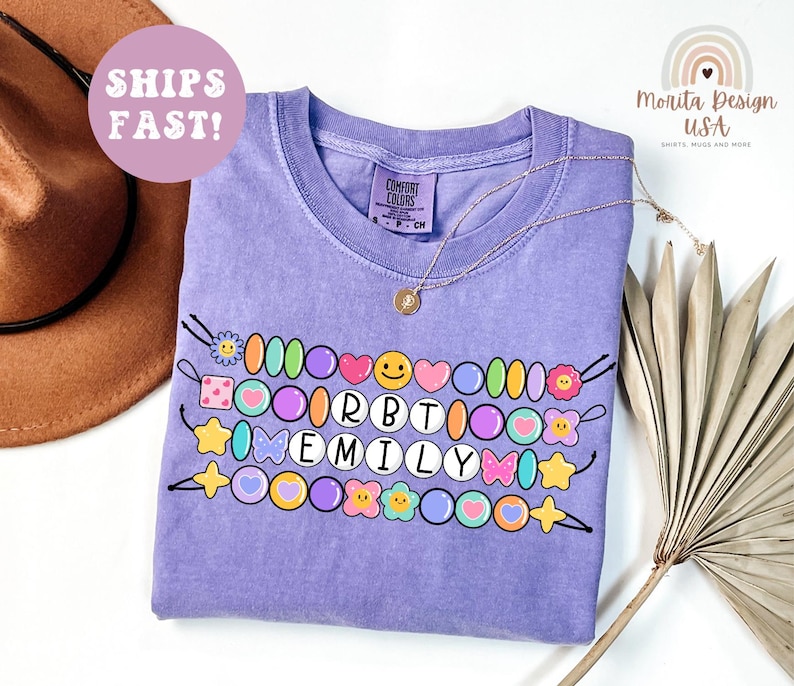May include: A light purple Comfort Colors t-shirt with a beaded design spelling "RBT EMILY". The design includes colorful beads, flowers, hearts, and smiley faces. A gold necklace and a brown hat are also visible.