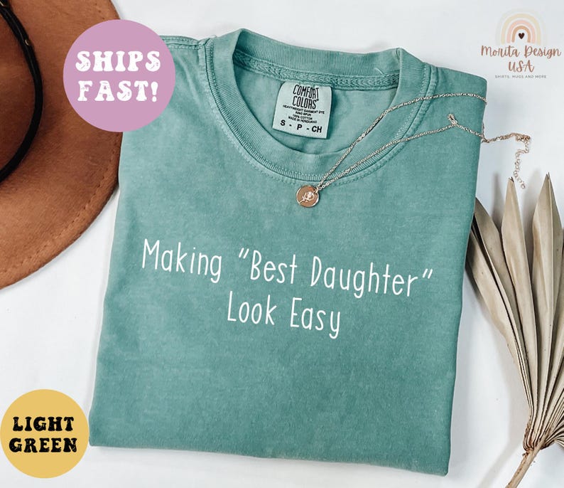 May include: A light green t-shirt with the text "Making "Best Daughter" Look Easy" in white. A gold necklace with a pendant rests on the shirt. The shirt has a Comfort Colors tag.
