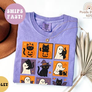 May include: Violet-colored t-shirt with a Halloween-themed graphic. The design features black cats, pumpkins, ghosts, and bats in orange and white squares. The shirt has a crew neck and the text "Comfort Colors" on the tag.