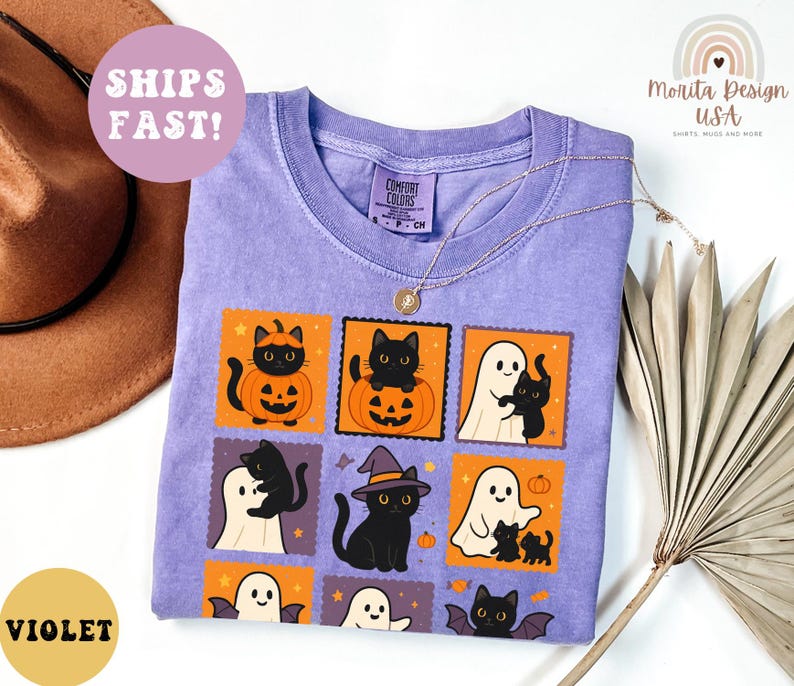 May include: Violet-colored t-shirt with a Halloween-themed graphic. The design features black cats, pumpkins, ghosts, and bats in orange and white squares. The shirt has a crew neck and the text "Comfort Colors" on the tag.