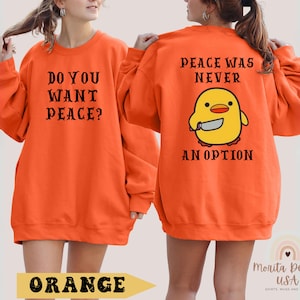 K&ouml;nnte beinhalten: Orangefarbener Pullover mit dem Text "Do you want peace?" auf der Vorderseite und "Peace was never an option" mit einem Cartoon-K&uuml;ken, das ein Messer h&auml;lt, auf der R&uuml;ckseite.