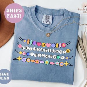 May include: Blue jean colored t-shirt with the words "OCCUPATIONAL THERAPY" in a colorful beaded design. The shirt has a crew neck and short sleeves. A gold necklace with a pendant is draped on the shirt. A round sticker says "SHIPS FAST!"