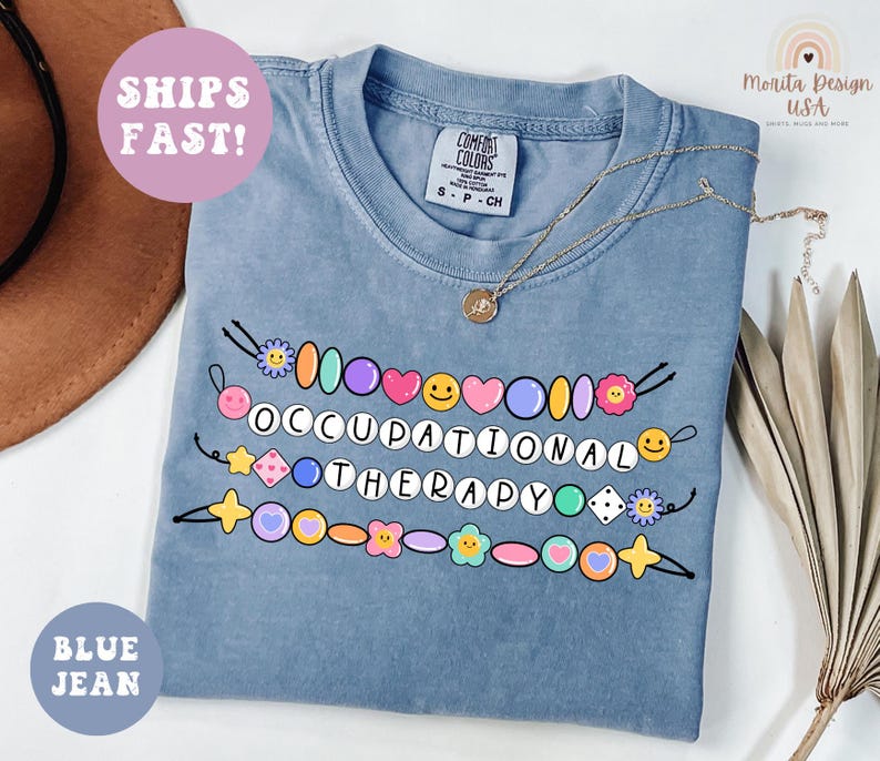 May include: Blue jean colored t-shirt with the words "OCCUPATIONAL THERAPY" in a colorful beaded design. The shirt has a crew neck and short sleeves. A gold necklace with a pendant is draped on the shirt. A round sticker says "SHIPS FAST!"
