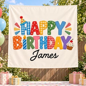 May include: A white fabric banner featuring the colorful text HAPPY BIRTHDAY in a stitched, multi-patterned style, with a small party hat on the H, a candle on the I, and a cupcake on the Y. The name James is printed in black script below.