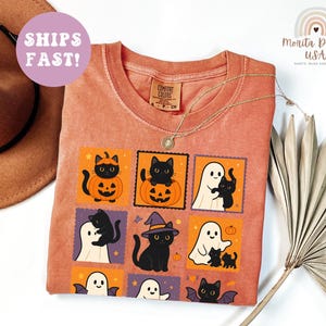 May include: Orange Comfort Colors t-shirt with a Halloween-themed graphic. The graphic features black cats, pumpkins, ghosts, and bats in a stamp-like design. A gold necklace is draped over the shirt. A brown hat is visible in the background.