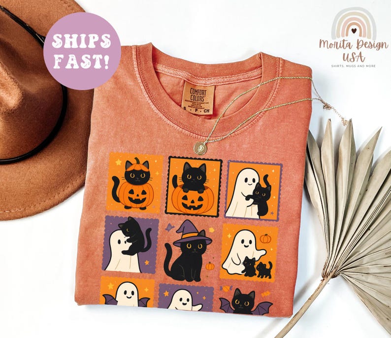 May include: Orange Comfort Colors t-shirt with a Halloween-themed graphic. The graphic features black cats, pumpkins, ghosts, and bats in a stamp-like design. A gold necklace is draped over the shirt. A brown hat is visible in the background.
