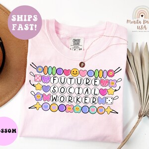 May include: A light pink t-shirt with the words "FUTURE SOCIAL WORKER" in a circle font. The shirt has colorful beaded accents and a necklace. The shirt also has the text "SHIPS FAST!" and "BLOSSOM".