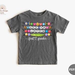 May include: Dark gray t-shirt with the words "SEE YA LATER" in colorful letters, along with various school-themed graphics. A green alligator is featured below the text, with the words "first grader" underneath.
