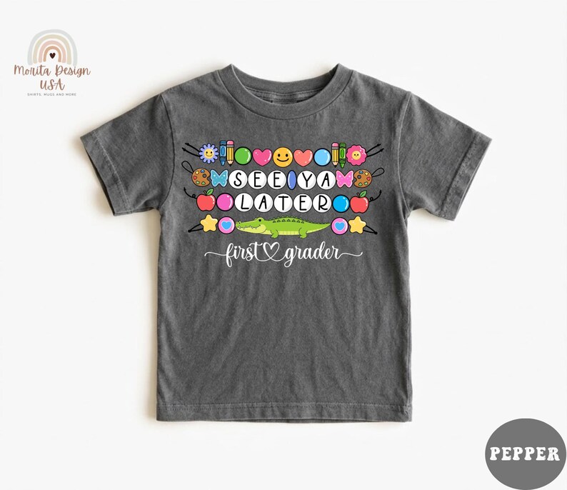 May include: Dark gray t-shirt with the words "SEE YA LATER" in colorful letters, along with various school-themed graphics. A green alligator is featured below the text, with the words "first grader" underneath.