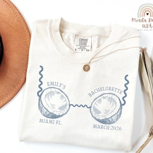 May include: Cream-colored t-shirt with a blue graphic of two coconuts connected by a wavy line, with the text "Emily's Bachelorette" and "Miami FL. March 2026". The shirt has a Comfort Colors tag and a gold necklace.
