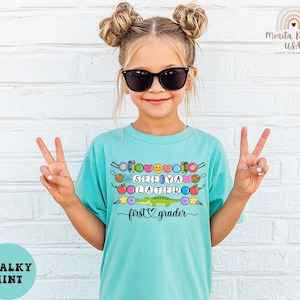 May include: A turquoise t-shirt with the words "SEE YA LATER" and "first grader" in a playful font. The shirt features colorful graphics of flowers, hearts, and a cartoon alligator. The girl is wearing sunglasses and making a peace sign.