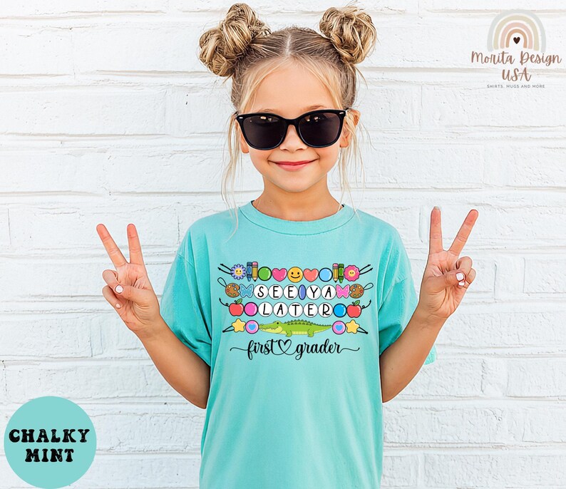 May include: A turquoise t-shirt with the words "SEE YA LATER" and "first grader" in a playful font. The shirt features colorful graphics of flowers, hearts, and a cartoon alligator. The girl is wearing sunglasses and making a peace sign.
