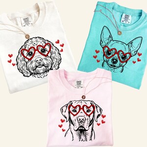 May include: Three Comfort Colors t-shirts in white, light blue, and pink. Each shirt features a dog graphic wearing heart-shaped glasses and surrounded by red hearts. A gold necklace with a pendant is draped on each shirt.