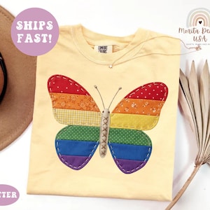 May include: A pale yellow t-shirt featuring a large butterfly graphic on the front. The butterfly wings are composed of horizontal fabric strips in red, orange, yellow, green, blue, and purple, with visible white stitching along the edges.