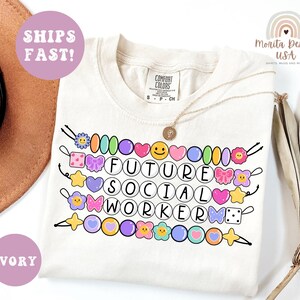 May include: Ivory-colored t-shirt with the text "FUTURE SOCIAL WORKER" in a circle font, decorated with colorful cartoon beads, hearts, and flowers. The shirt also has the text "SHIPS FAST!" and "IVORY" in a circle.