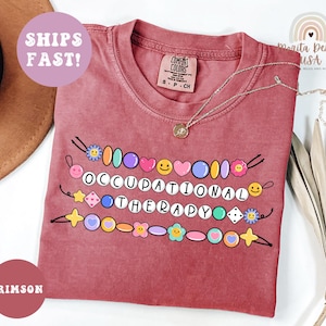 May include: Crimson-colored t-shirt with the words "OCCUPATIONAL THERAPY" spelled out with colorful beads and smiley faces. The shirt has a Comfort Colors tag and a silver necklace. The words "SHIPS FAST!" are in a circle.