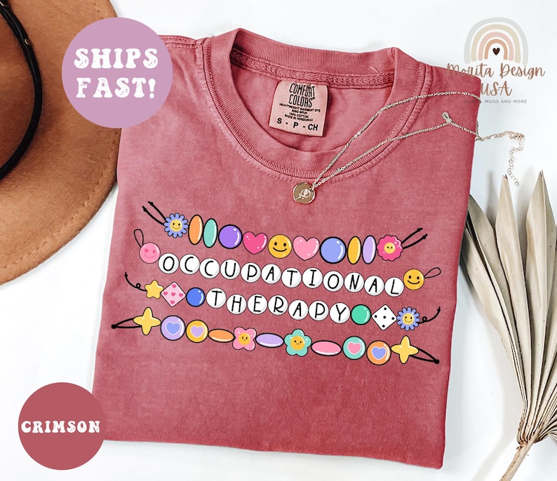 May include: Crimson-colored t-shirt with the words "OCCUPATIONAL THERAPY" spelled out with colorful beads and smiley faces. The shirt has a Comfort Colors tag and a silver necklace. The words "SHIPS FAST!" are in a circle.