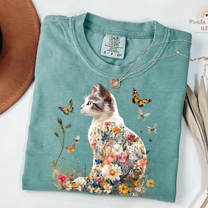 May include: A teal blue t-shirt featuring a white cat with flowers growing from its body. The cat is looking to the left and there are butterflies flying around it. The shirt has a tag that says "Comfort Colors" and "S - P - CH".