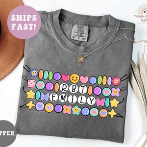 May include: A gray Comfort Colors t-shirt with a colorful beaded design that spells out "RBT EMILY". The design includes various shapes and colors, such as hearts, stars, and flowers. A gold necklace is draped across the shirt.
